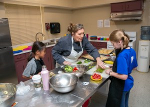Alt text -- An adult instructs two children in a kitchen setting.